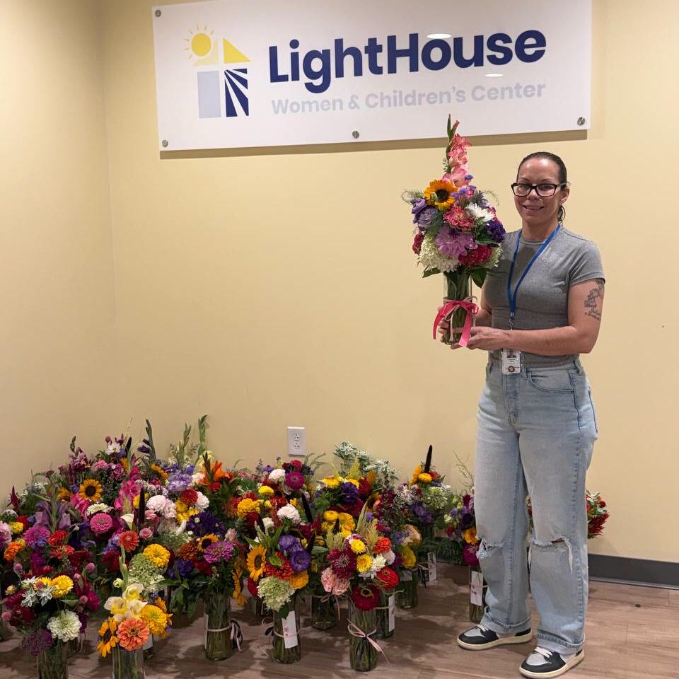 A staff member holds a bouquet beneath the LightHouse Women & Children’s Center sign, with rows of filled vases lined up along the wall behind her.