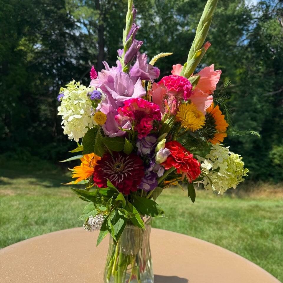 A finished bouquet of gladiolus, hydrangea, dahlias, celosia and a sunflower in a glass vase on an outdoor table.