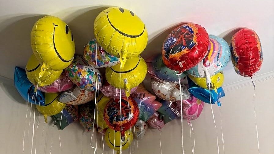 A ceiling full of smiley face, Spider-Man, unicorn and confetti balloons, their ribbons hanging down the wall.