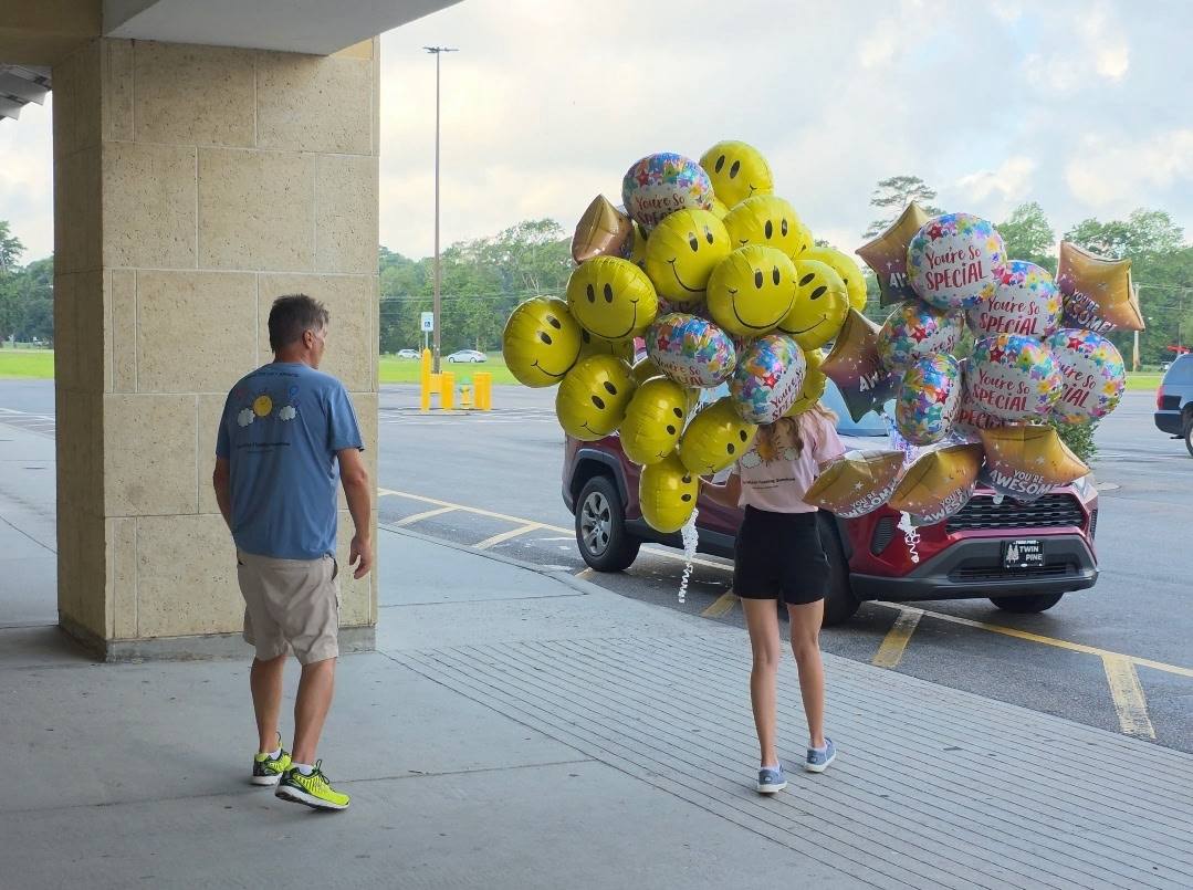 A big bundle of smiley face, star, and You're So Special balloons, all blown up and ready for delivery, is carried to a car in a parking lot.