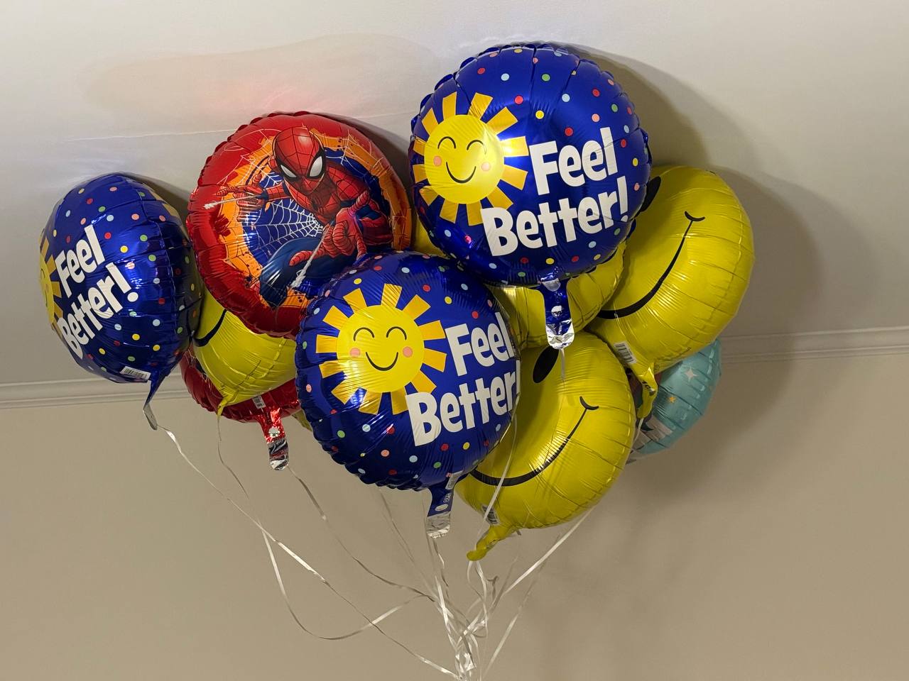 A bouquet of foil balloons floats against a ceiling, including Feel Better sunshine balloons, yellow smiley faces, and a Spider-Man balloon.