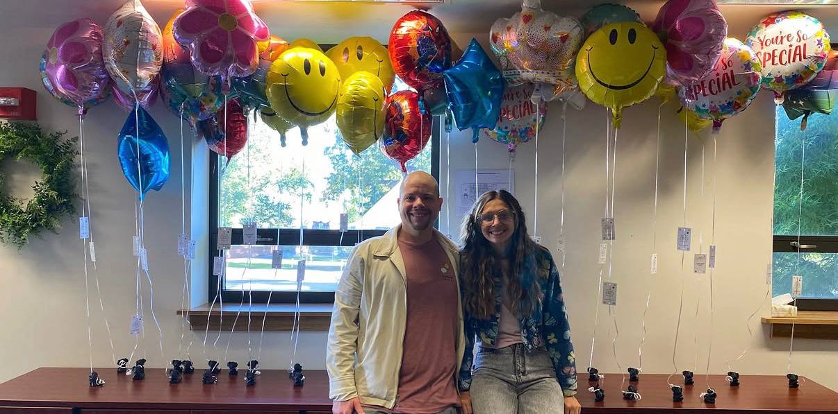 Two volunteers smile in front of a long row of balloons, each tied to a small 3-D printed weight shaped like Teddy the dog.
