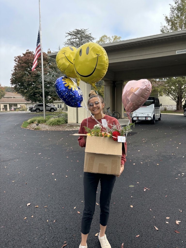 Ashton stands outside the retirement community holding a cardboard box of bouquets with smiley face and heart balloons floating above it.